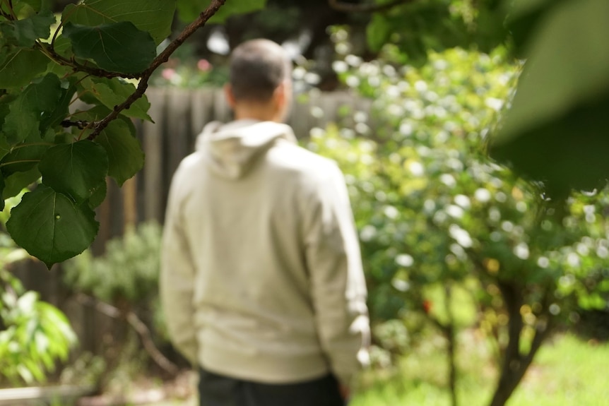 A man in a beige hoodie stands in a garden, out of focus, with his back to the camera.