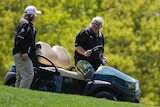 A golfer talks to a man as he gets into a golf cart on a grassy slope.