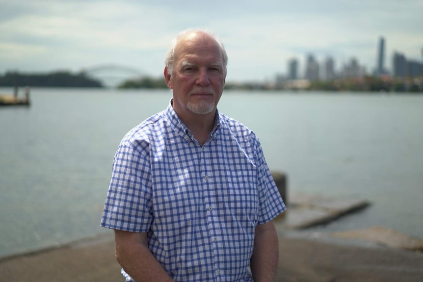 An older man in a blue and white check shirt with a view of the harbour behind him.