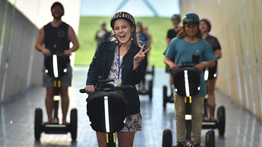 A group of young people on segways roll toward the camera laughing outside parliament house.