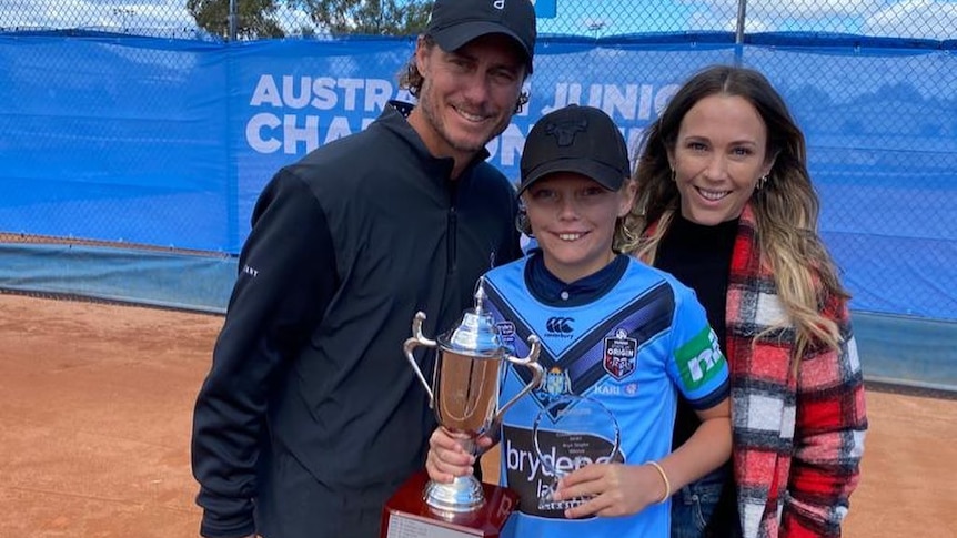 A junior male tennis player holding trophies as he stands alongside his parents.