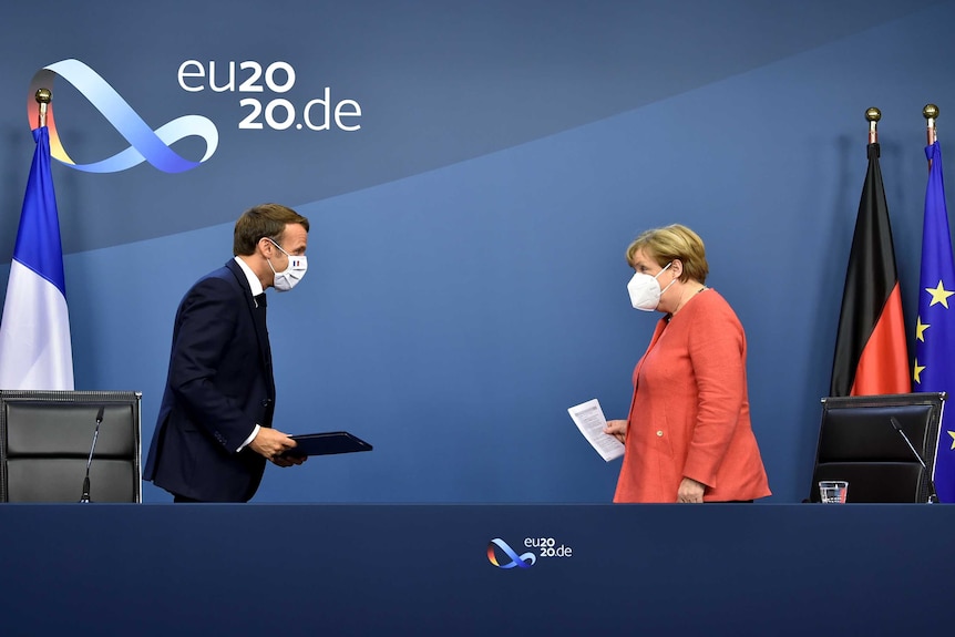 A middle-aged man looks towards an older woman, both wearing masks, on a blue stage.