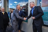 Two men with greying hair in suits shake hands on a street watched by other people