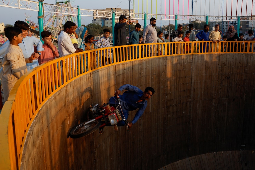 An excited crowd watches a rider with no helmet speeding along a circular timber wall on a motorbike.
