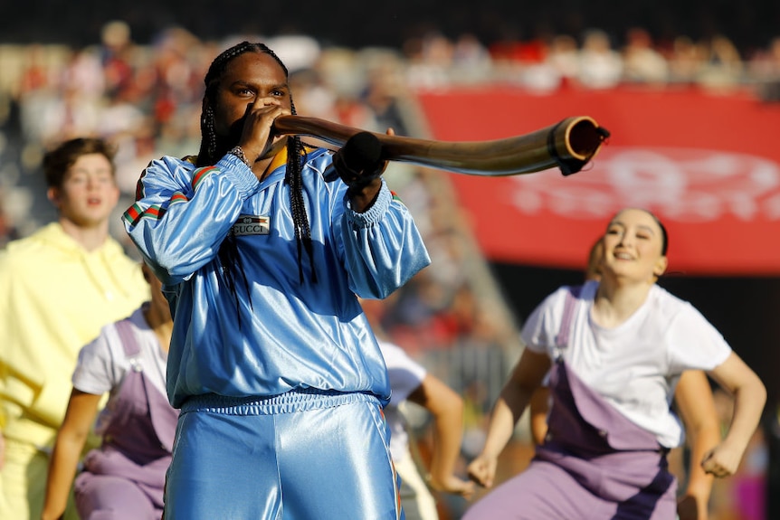 artist baker boy plays a digeridoo while wearing a baby blye satin gucci tracksuit. there are backup dancers behind him