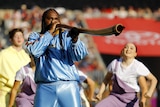 artist baker boy plays a digeridoo while wearing a baby blye satin gucci tracksuit. there are backup dancers behind him