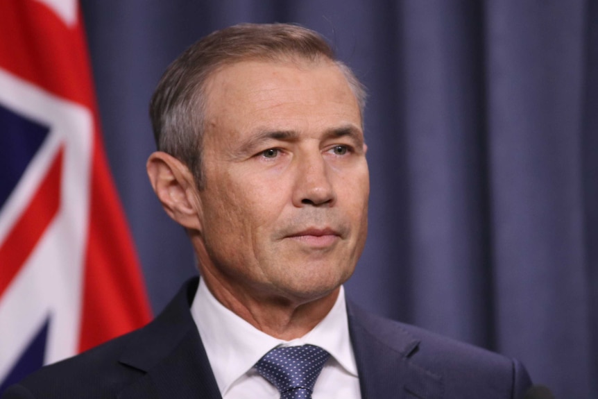 A tight head and shoulders shot of WA Health Minister Roger Cook indoors with a flag behind him.