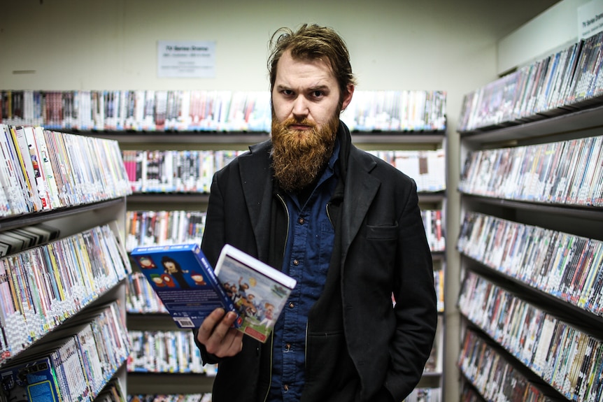 Lloyd Dodsworth holding two DVDs in his hand against a backdrop of shelves filled with DVDs.