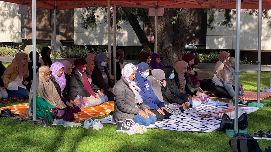 A group of Islamic women gathered under a tent on a lawn for midday prayers.