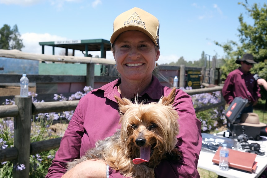 A woman wearing a pink collared shirt and holding a dog smiles at the camera.
