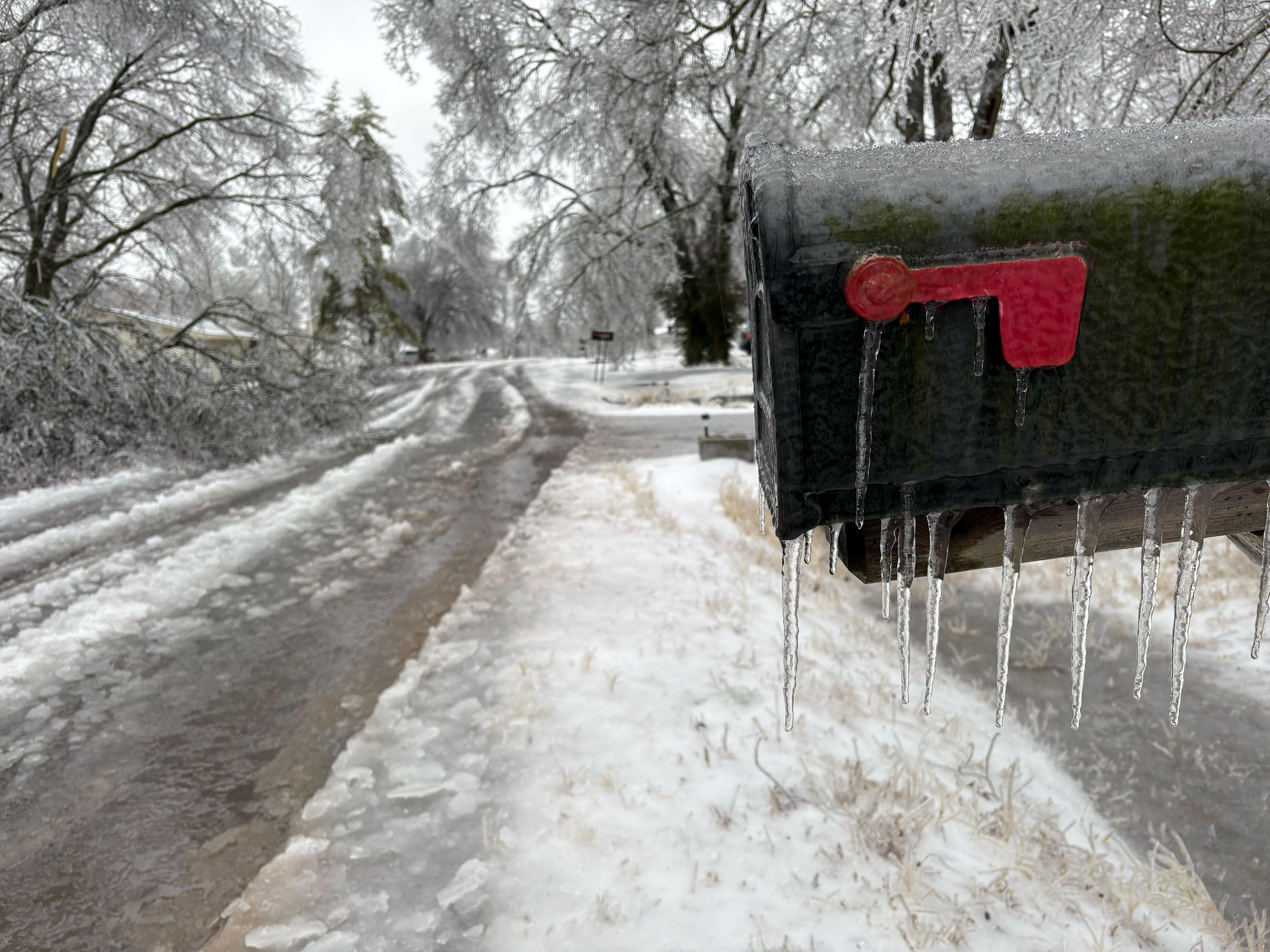 Carámbanos cuelgan de un buzón con una calle sucia, helada y nevada al fondo y árboles cubiertos de nieve.