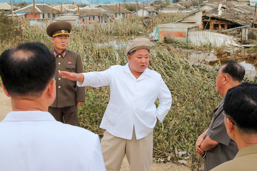 Kim Jong-un in a beige hat talking to a grow of men in a village damaged by a typhoon