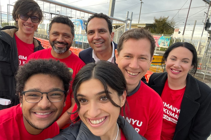 A group of people wearing red 'Abi Kumar' t-shirts pose for a photo at a train station