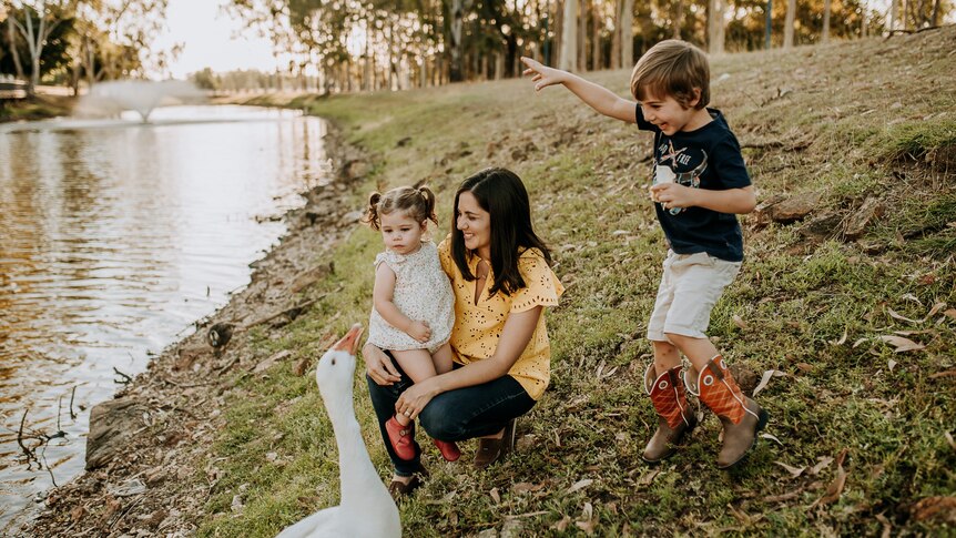 A woman and her children are near a pond. One child on the woman's lap, an older child is jumping, a goose is in the foreground.