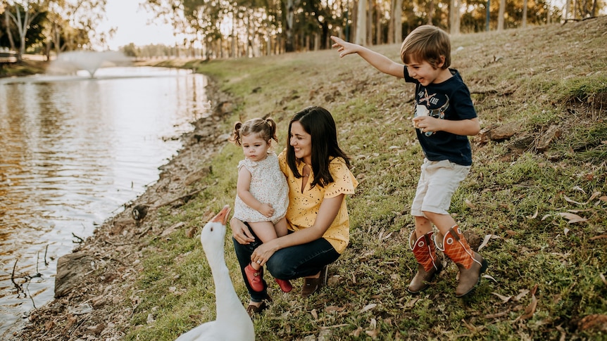 A woman and her children are near a pond. One child on the woman's lap, an older child is jumping, a goose is in the foreground.