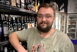 a man with brown facial hair leaning against a row of wine bottles