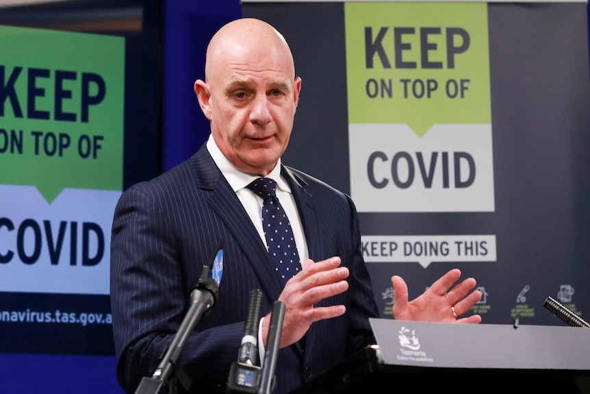 Tasmanian Prime Minister Peter Gutwain signals in front of a sign "Keep COVID on top".