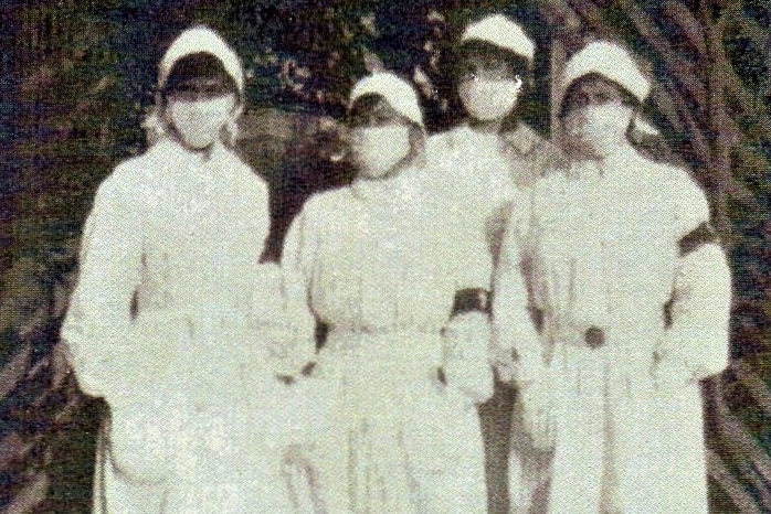 A black and white photo showing four nurses wearing masks and gowns.