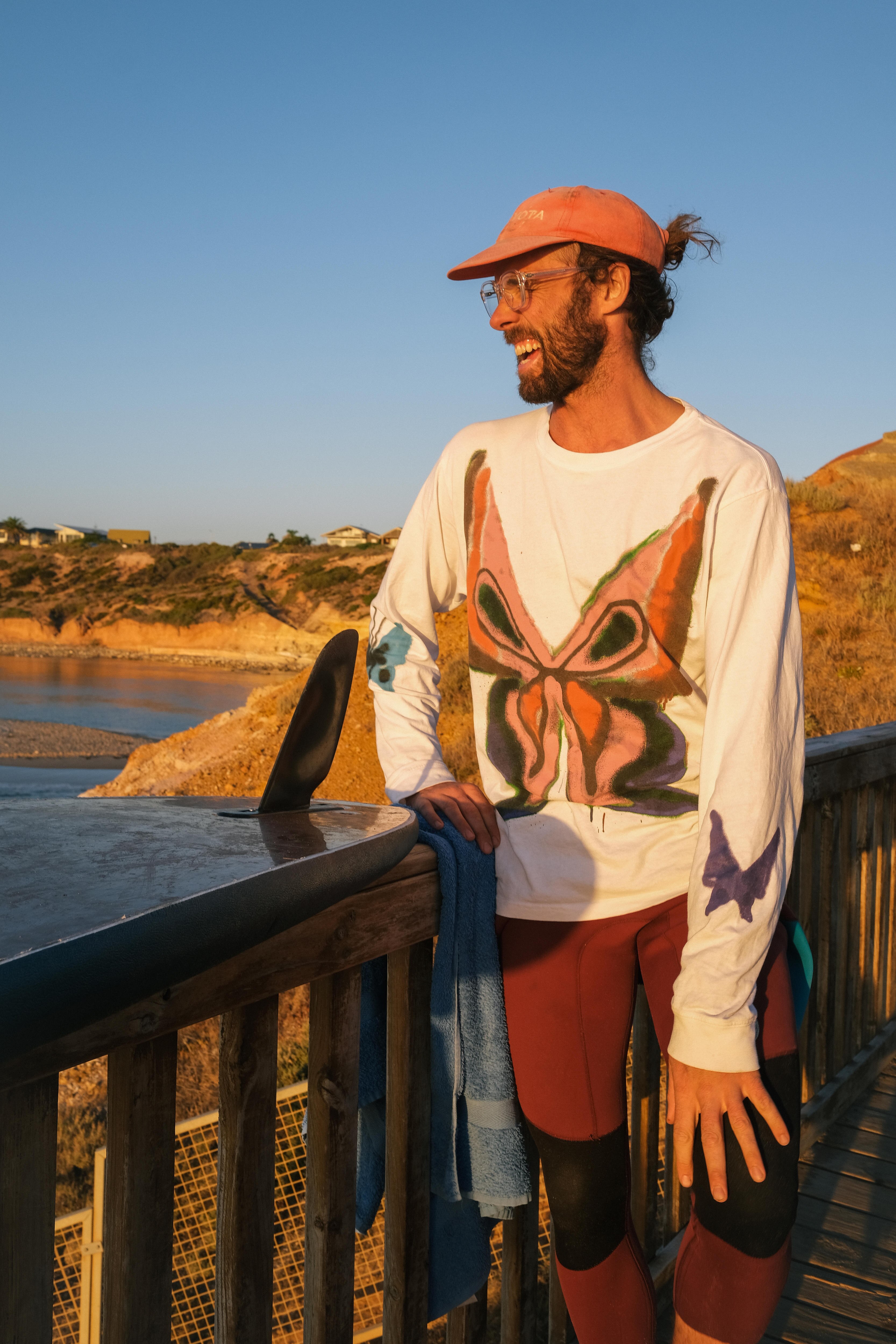 Man in red hat and tshirt with butterfly on it laughs next to the ocean