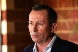 WA Premier Mark McGowan wearing a white check shirt and jacket standing in front of a brick wall.