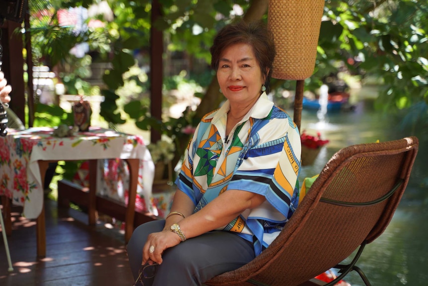 A woman with brown hair wears a colourful shirt while she sits on a chair surrounded by trees.