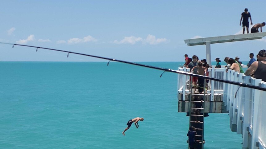 A teenager jumps off into the blue water, while others wait on the roof of the shad structure