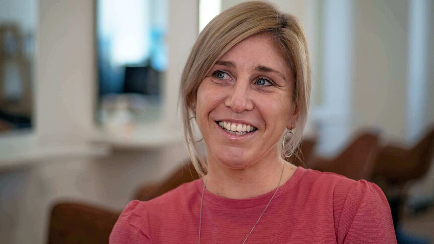 a close up portrait of a blonde haired smiling woman, in a pink top