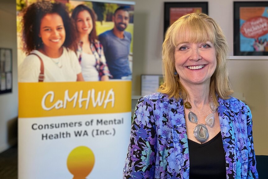 A smiling woman with blonde hair poses for a photo indoors in front of a Consumers of Mental Health WA poster.