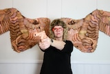 A woman stands holding a gun made of plastic hands in front of wings made from doll parts