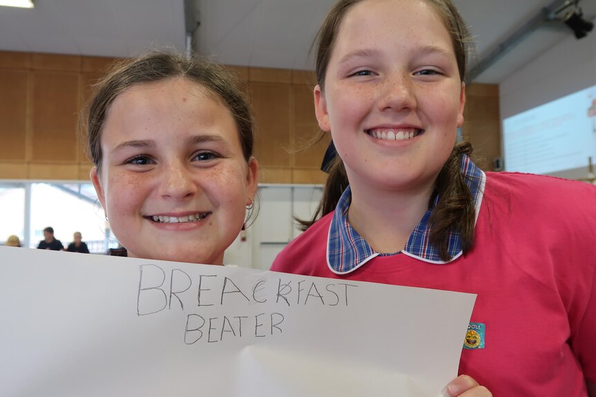 Two smiling schoolgirls hold a large sheet of paper with the handwritten words breakfast beater.  