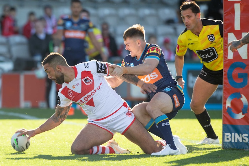 Paul Vaughan grounds the ball with his right hand. A Titans player is holding onto his left arm. A referee watches on closely.