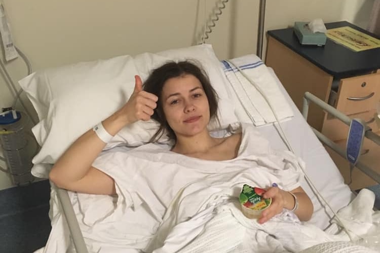 Bridget Hustwaite is in a hospital bed wearing a white gown with a tub of food in her hand. She's putting one thumb up.