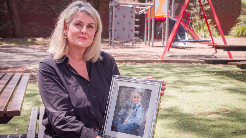 A woman holding a photograph of her young son in a park.