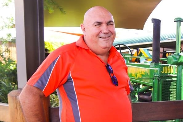A bald, middle-aged man wearing a red shirt is smiling with his hands on his hips. His sunglasses are tucked into his shirt