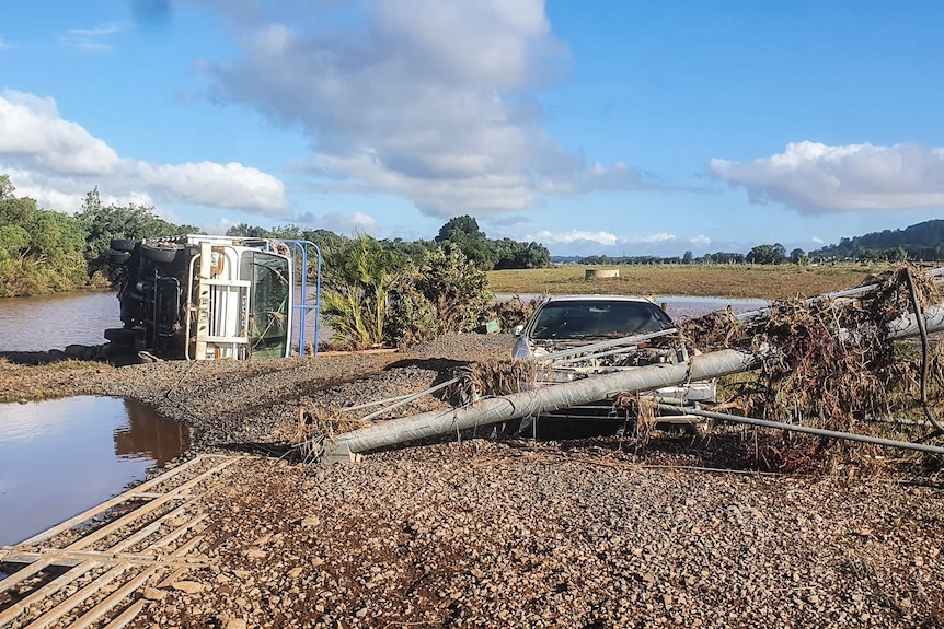 Truck on its side, car covered with debrief next to large piles of flood water.