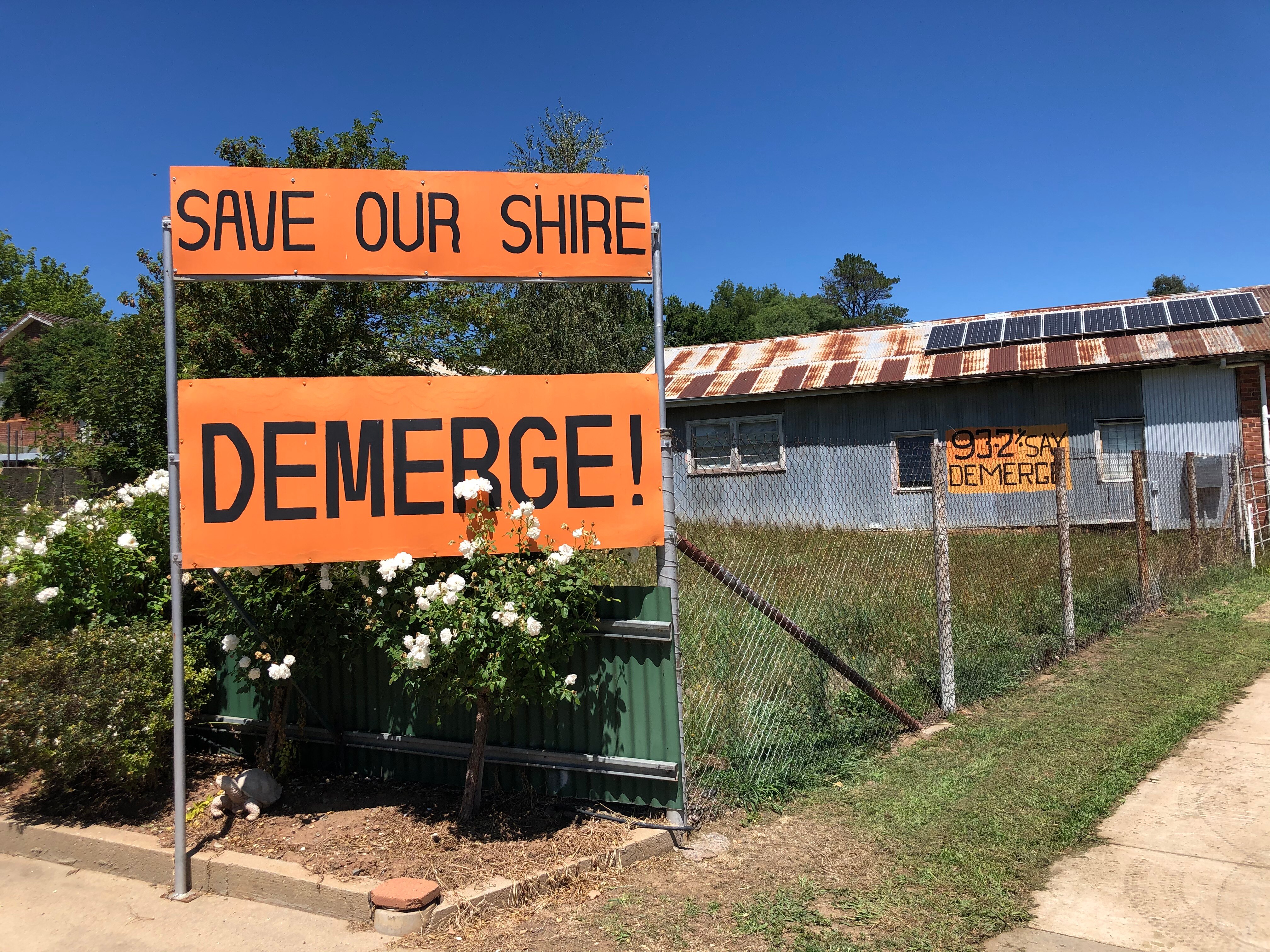 Orange signs read "save our shire" "DEMERGE!" on a fence line
