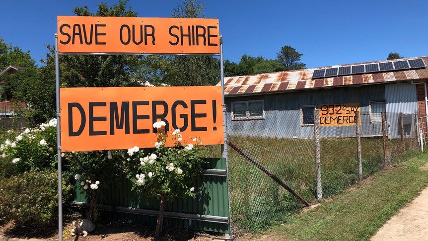Orange signs read "save our shire" "DEMERGE!" on a fence line