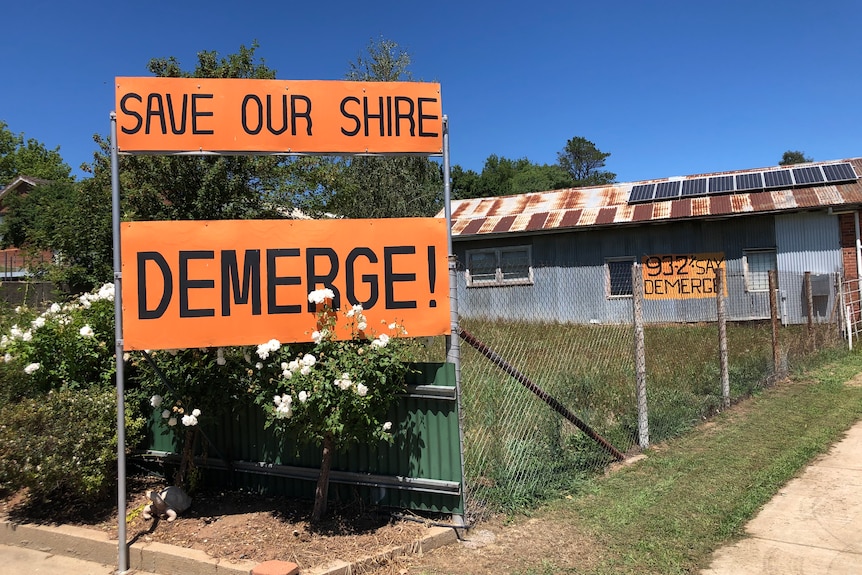 Orange signs read "save our shire" "DEMERGE!" on a fence line