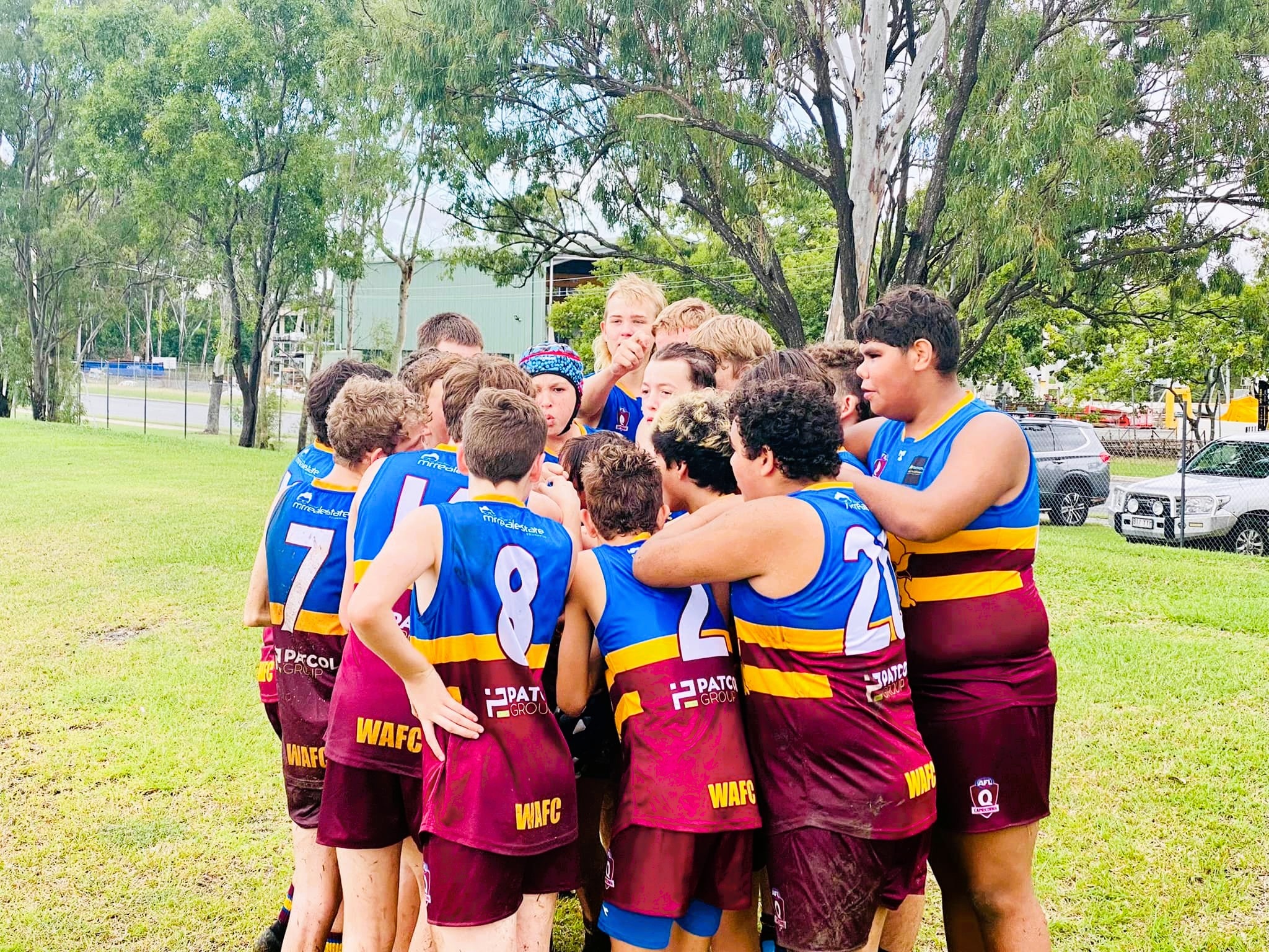A group of Aussie Rules junior players in maroon, yellow and blue singlets in a huddle at a field.