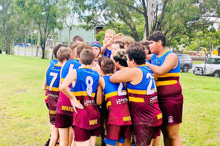 A group of Aussie Rules junior players in maroon, yellow and blue singlets in a huddle at a field.