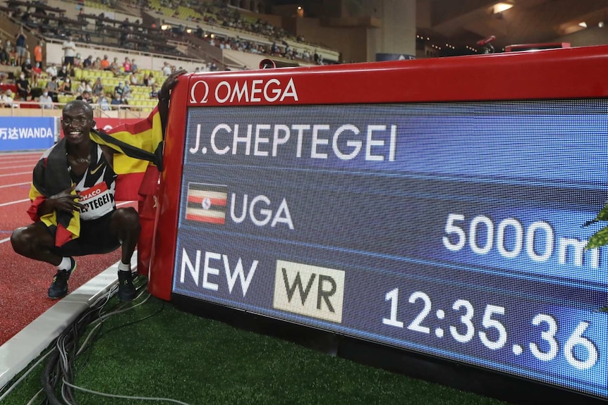 Joshua Cheptegei with a flag wrapped around him squats on the running track next to the timing board showing his race time