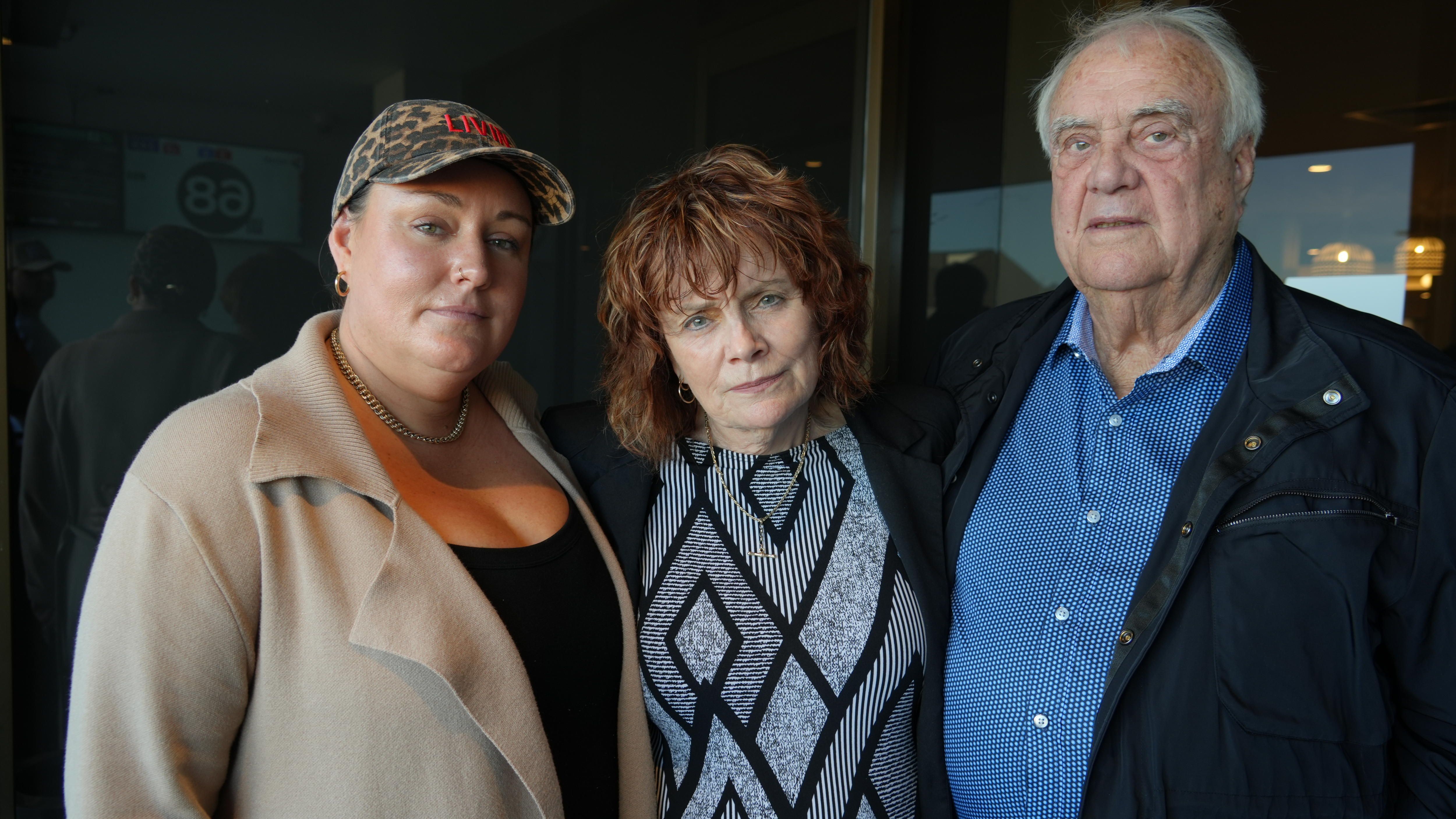Three sombre people, two women and a man, standing arm in arm.