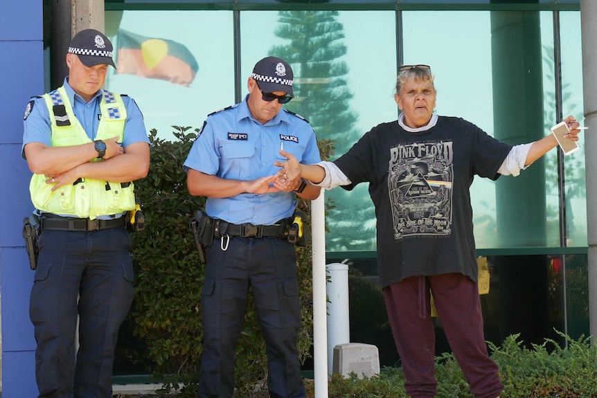 An Aboriginal woman stands outside a police station with her arms outstretched next to two officers looking down to the ground.