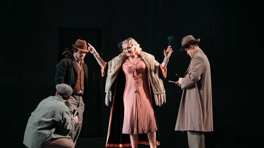 On stage a woman in 1920s formal outfit is surrounded by men