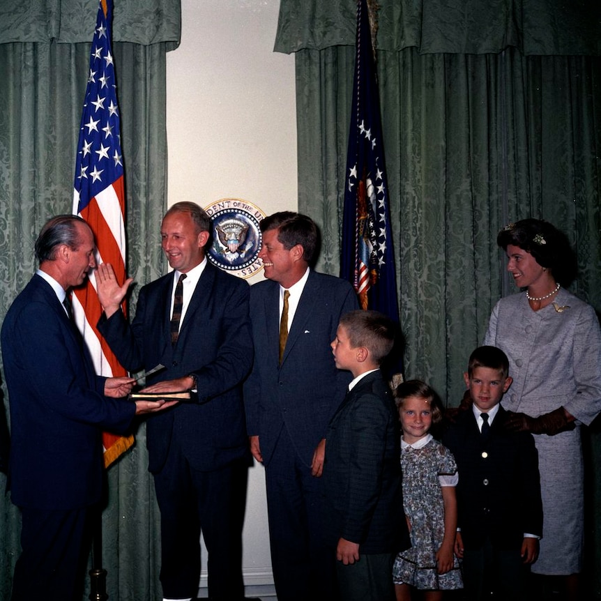 JFK grinning while a man holds his hand on a bible and his other hand up in the air 