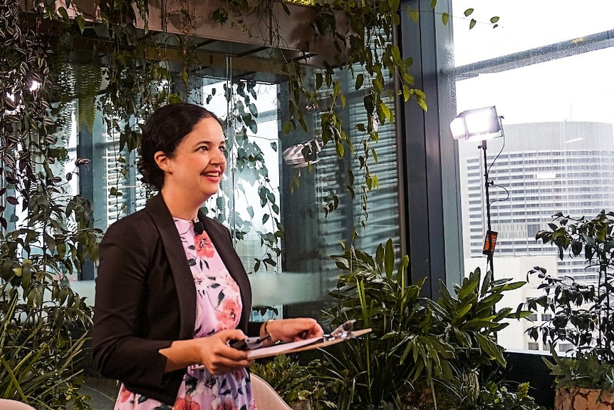 Davina Rooney holds a clipboard and smiles in a room filled with plants.