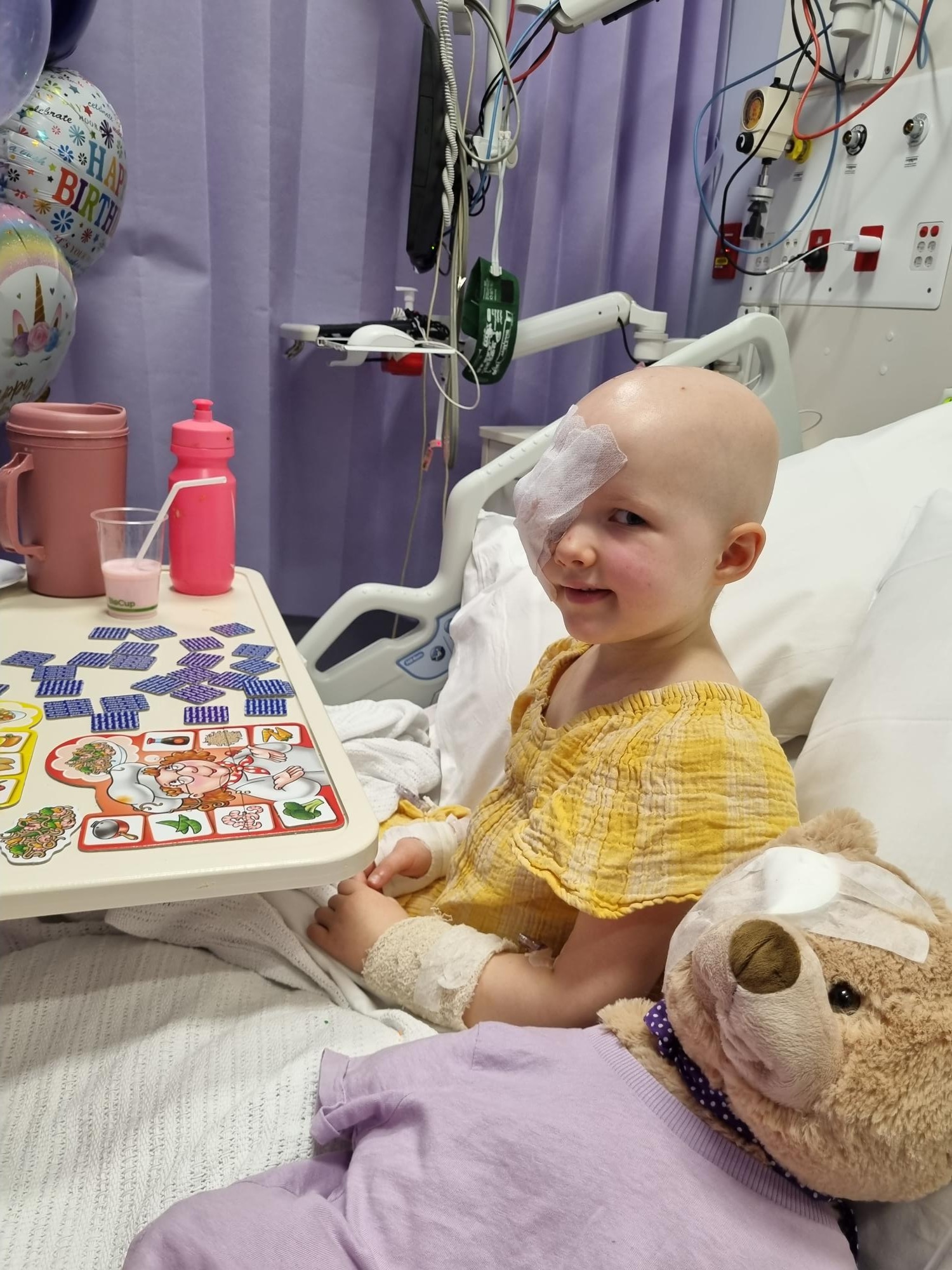 Alice sitting in a hospital bed, eye patch on, teddy bear in front, games on a table in front of her.