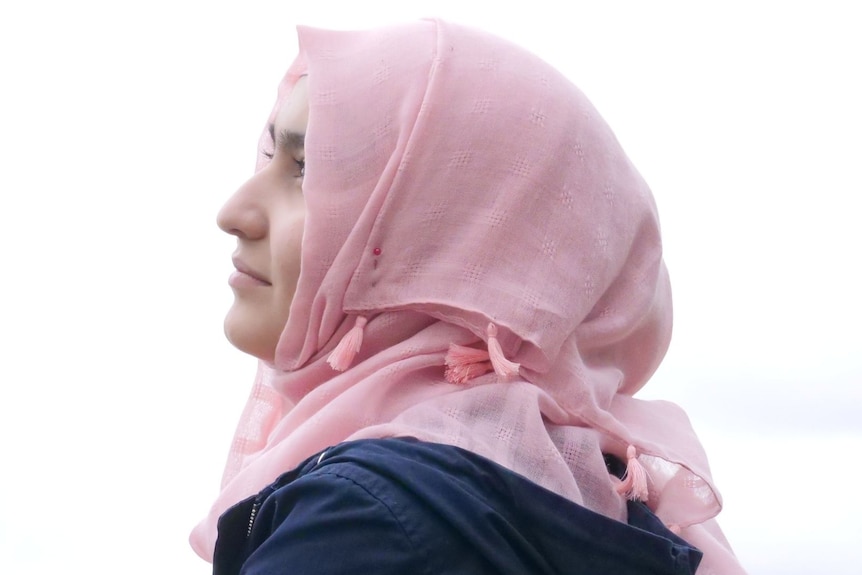 A teenage girl wearing a pink hijab looks to the side of the camera. Half her face is visible. 