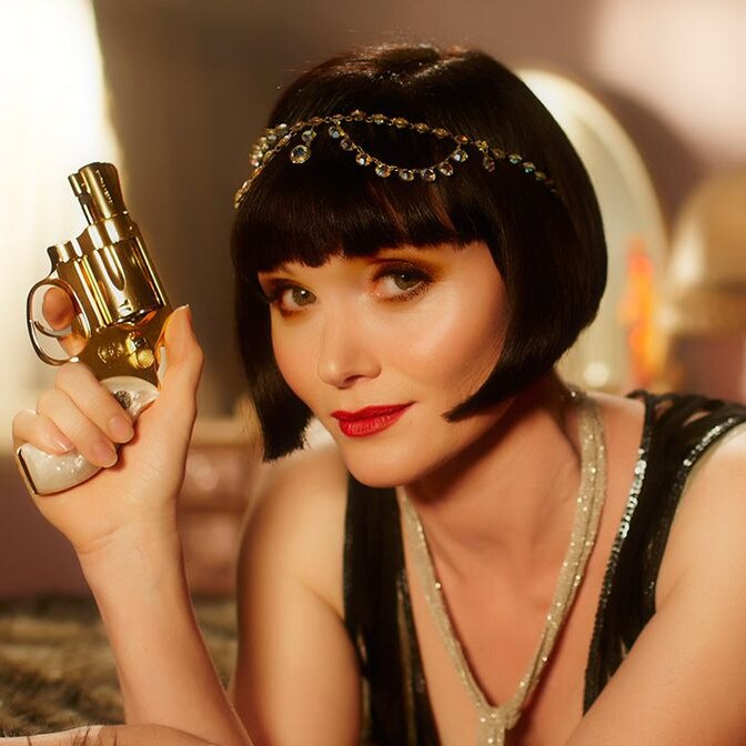 Actor Essie Davis in 1920s flapper costume, lying on a bed holding a pistol while looking suggestively at the camera.