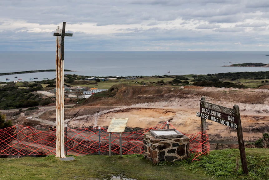 A monument with a Christian cross on a pole sits on a cliff edge overlooking a large open cut mine with ocean in background